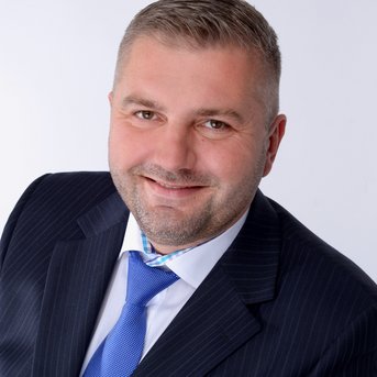 Mann mit kurzem, grauem Haar, trägt einen Anzug mit Krawatte und lächelt freundlich in die Kamera. Hintergrund ist neutral.