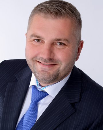 Mann mit kurzem, grauem Haar, trägt einen Anzug mit Krawatte und lächelt freundlich in die Kamera. Hintergrund ist neutral.